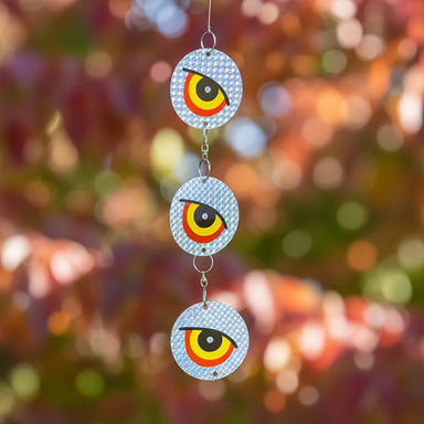 Three circular hanging wind spinner ornaments with a stylized red, yellow, black, and white eye design, displayed outdoors against a background of autumn foliage.